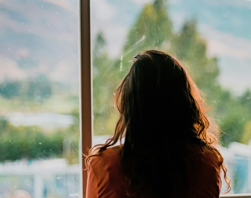 image of the back of a woman, looking reflectively outside the scenic view through the window, lost in her thought.