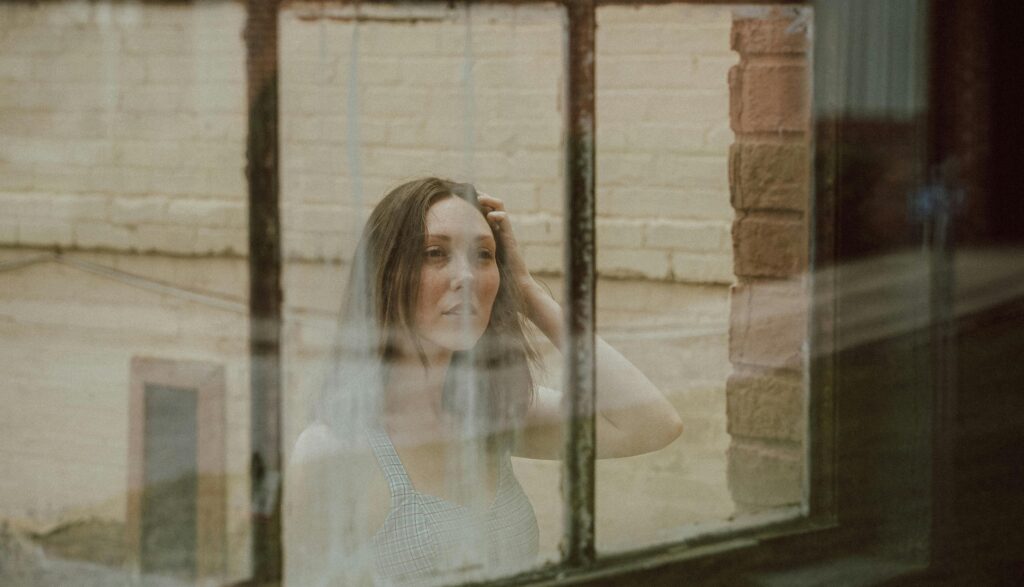 A woman looks through a vintage window with her hand in her hair, appearing pensive.