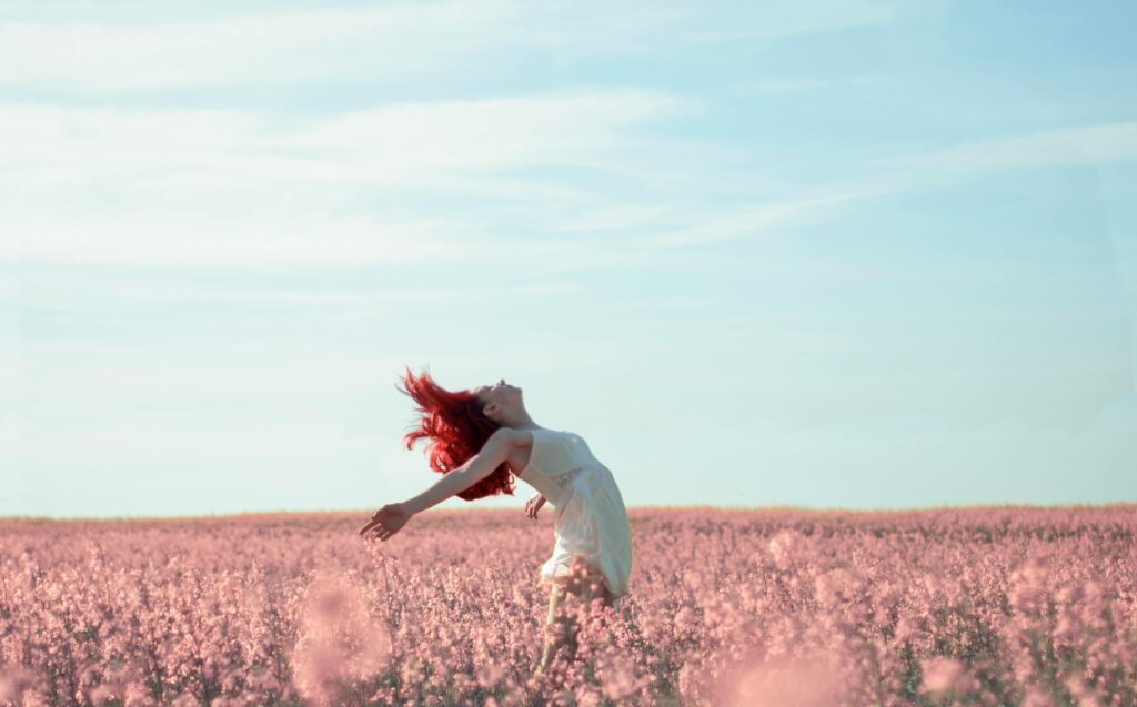 A woman with red hair wearing a white dress leans back with arms outstretched in a vast field of pink flowers under a clear blue sky. Schizoaffective disorder vs schizophrenia.