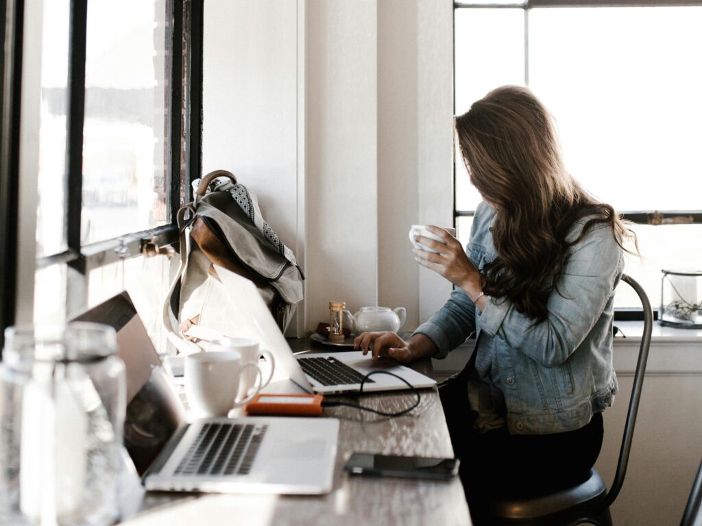 A woman sitting by a window in a cafe, holding a cup of coffee and working on her laptop, with a backpack resting nearby, illustrating potential time blindness symptoms as she focuses intently on her tasks.